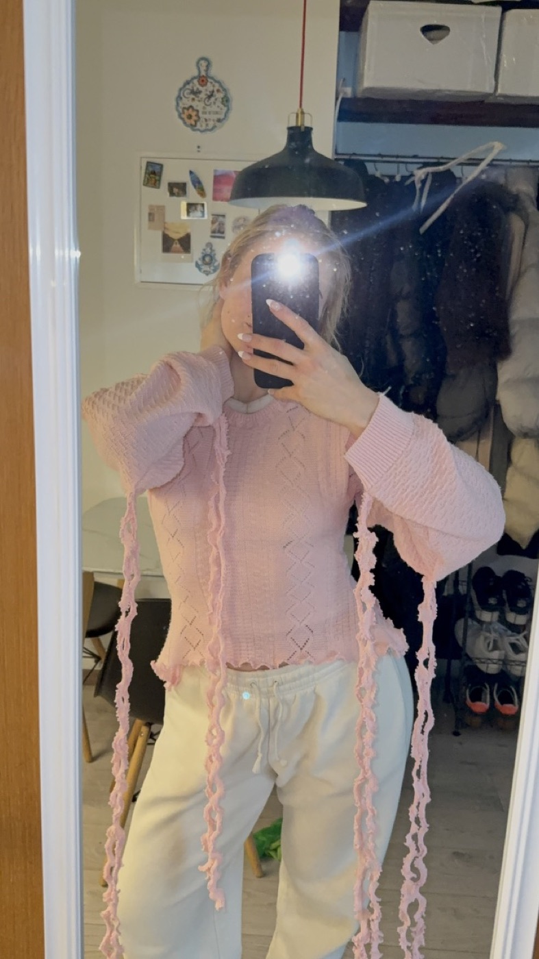 Pale pink knit top with long ruffled ribbon ties trailing from the sleeves
